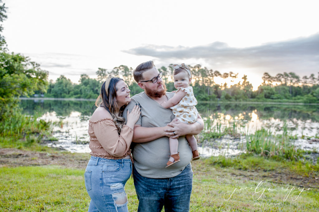 Mom leaning up against Dad who is holding infant daughter standing in front of a lake at sunrise. 