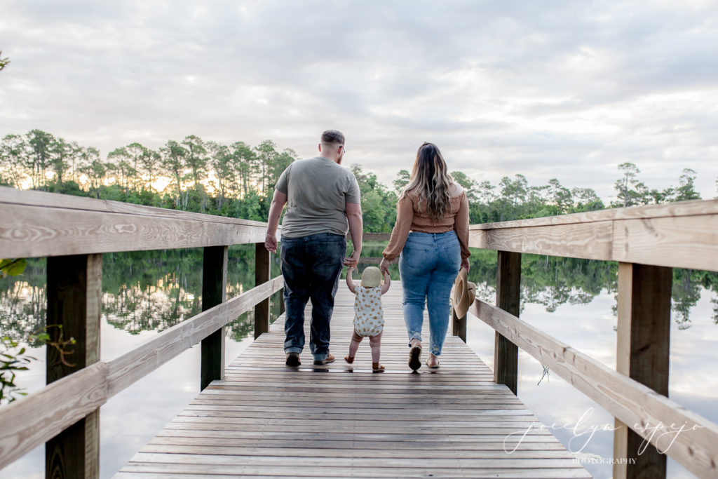 Mom and Dad holding baby girls hand as they walk down a dock.