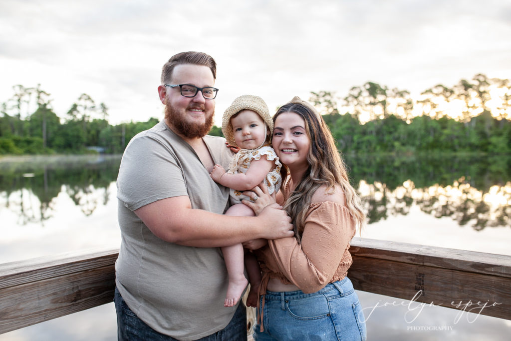 Mom and Dad holding baby at the corner of a dock on a lake. 