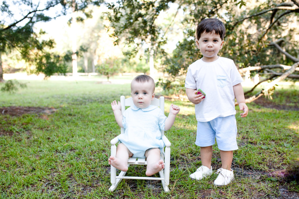 Baby boy sitting in tiny rocker with toddler brother standing next to him.