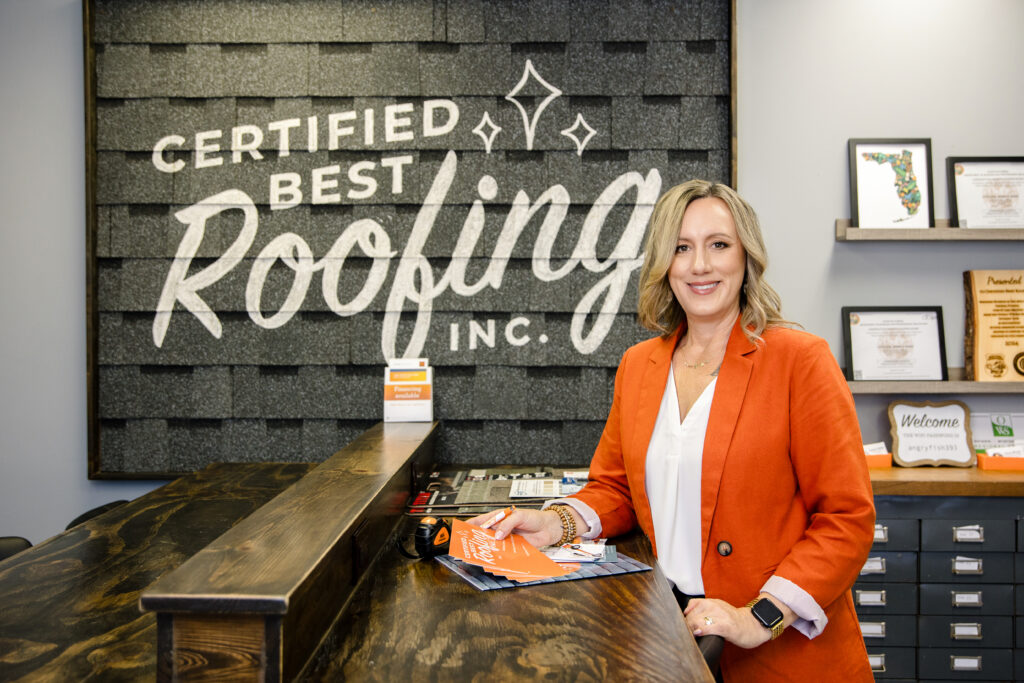 Lifestyle brand photo Female business owner standing confidently at the front desk of Certified Best Roofing, wearing an orange blazer and smiling in a modern office space by Branding by Jo.
