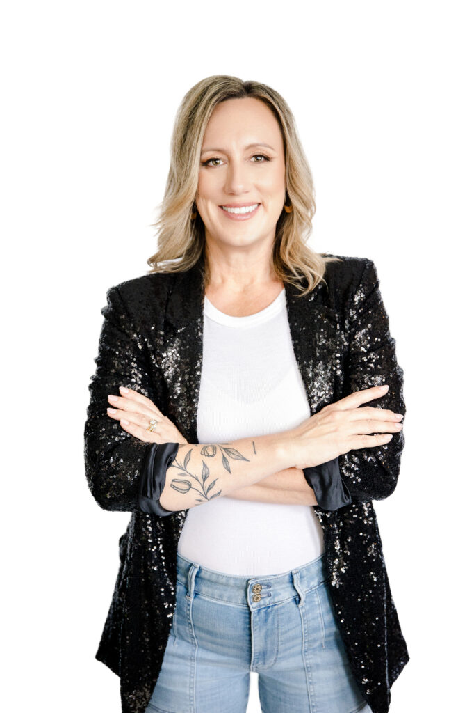 Confident female founder with blonde hair, dressed in a black sequin blazer, white blouse, and light blue jeans, smiling with arms crossed against a clean white background. Captured by Branding by Jo.