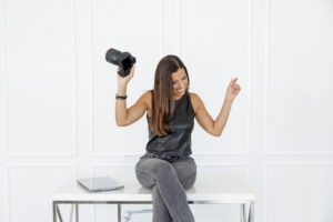 Woman in a black top and jeans sits on a white bench, playfully holding a camera and celebrating against a plain studio backdrop.