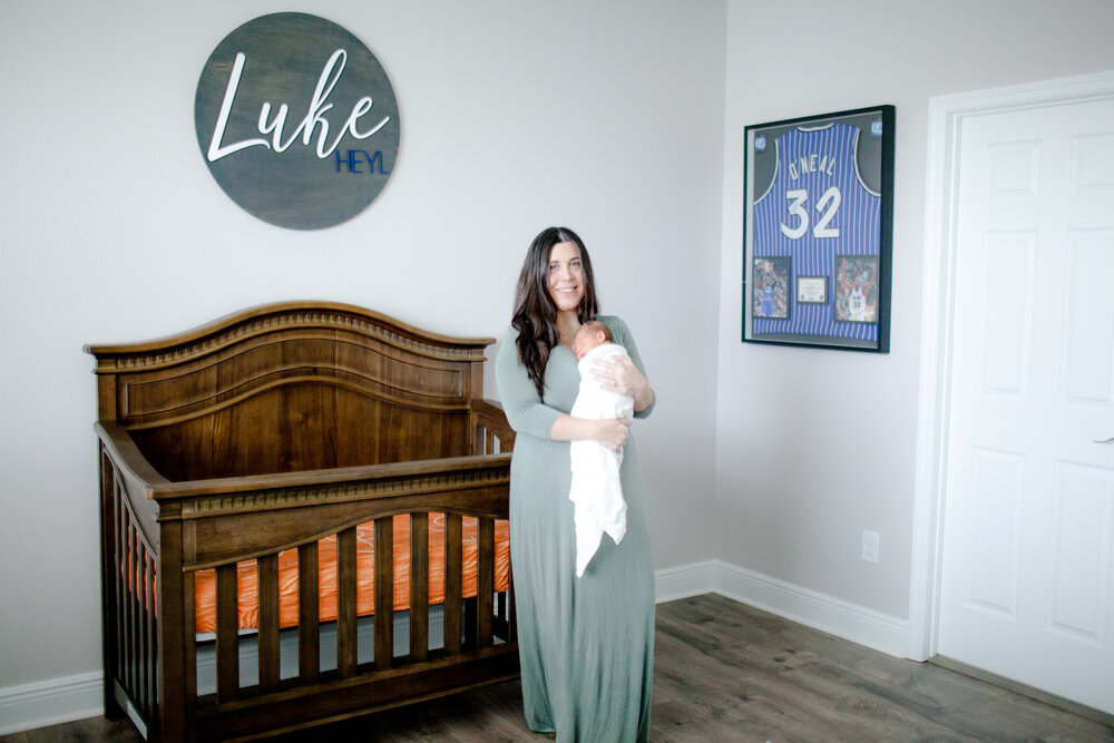 Oviedo, FL Mom in green flowy dress with newborn in white swaddle standing in front of crib in nursery decorated with basketball jersey and baby's name on the wall.