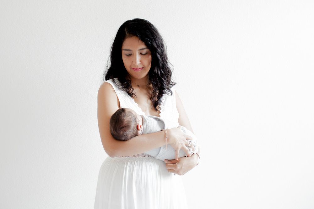 Mom standing with newborn baby swaddled in her arms, wearing a white flowy dress.  White background with natural light.