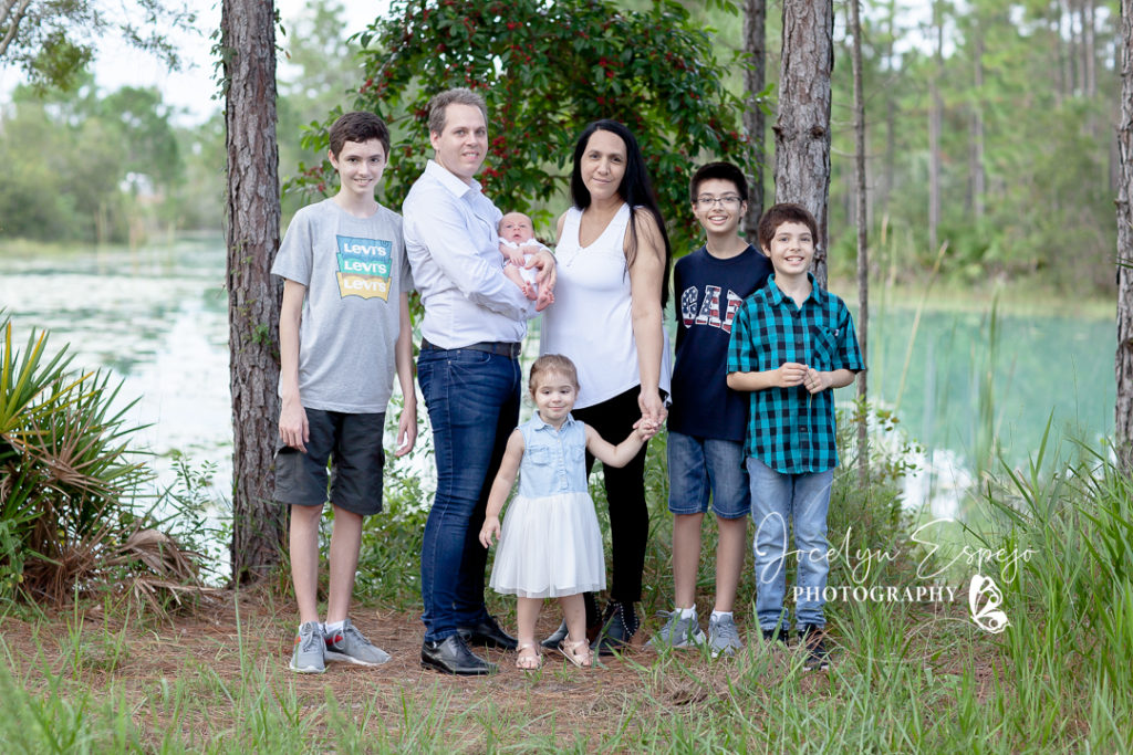 Outdoor lifestyle family photograph in front of a lake.  Mom and dad with their three older boys, toddler little girl and newborn baby boy.