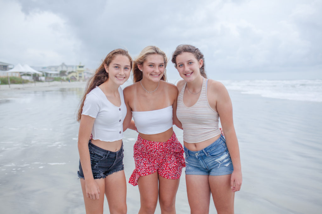 Three Girl best friends arms wrapped behind their backs smiling at the camera, New Symrna Beach in the background.