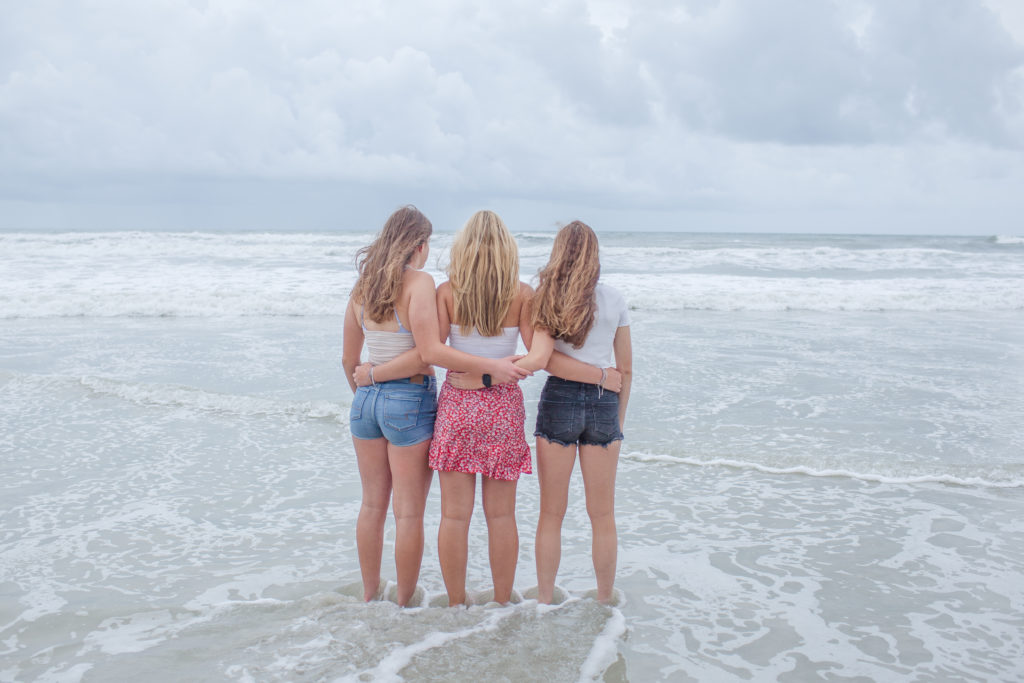 Three Girl best friends arms wrapped behind their backs facing away from the camera looking at the ocean in New Symrna Beach 