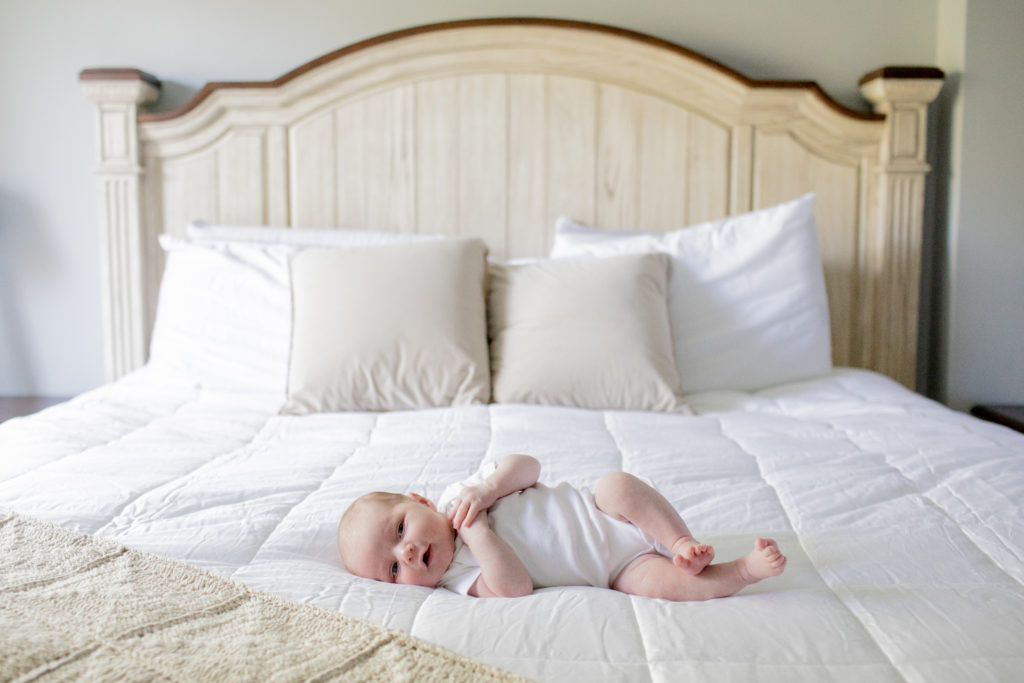 Newborn baby in white onesie on king bed with white blanket and wood headboard.
