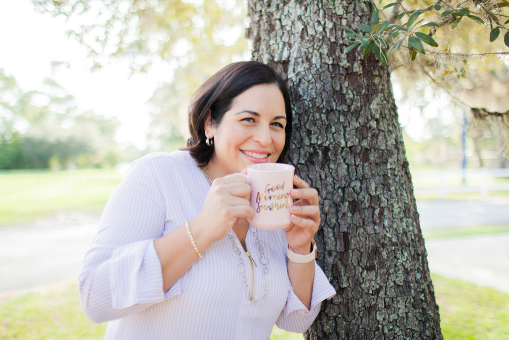 Women in a blush top holding a blush coffee mug smiling by a tree