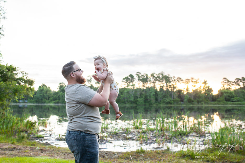 Dad holding baby girl up in front of his face at sunrise by a lake. 