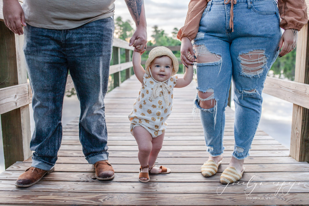 Baby girl standing on a dock holding mom and dad's hands and all you can see is their legs.