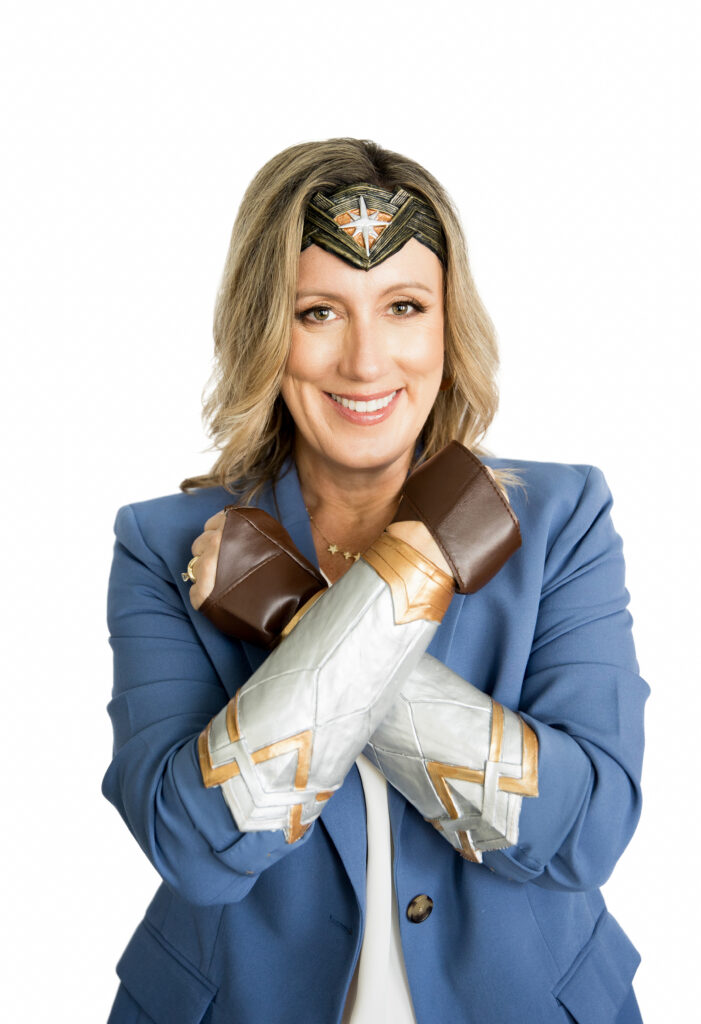 Smiling woman with blonde hair wearing a blue blazer and Wonder Woman headpiece and cuffs, standing with arms crossed in a power pose against a white background. Brand photo by Branding by Jo.
