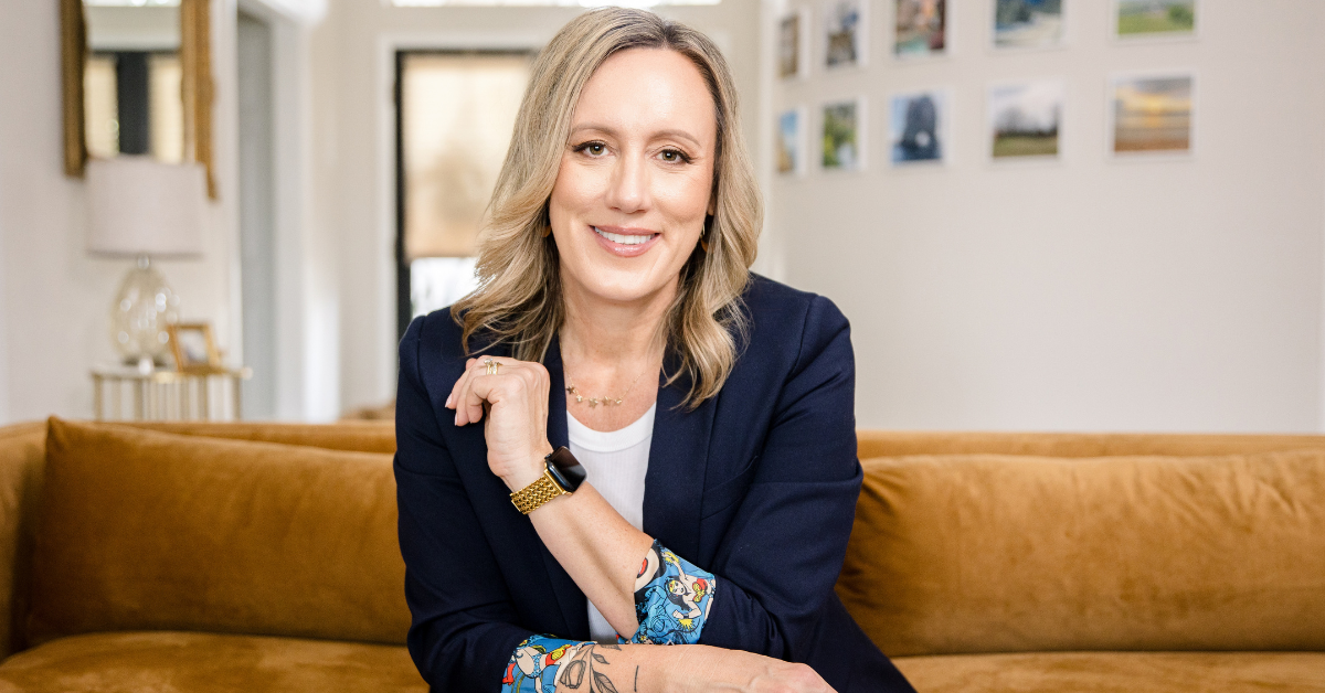 Confident woman with blonde hair in a navy blazer, smiling warmly while seated on a golden velvet couch, with Wonder Woman print details on her blazer sleeves and tattoos visible on her forearm. Photographed in a modern, light-filled living space by Branding by Jo.