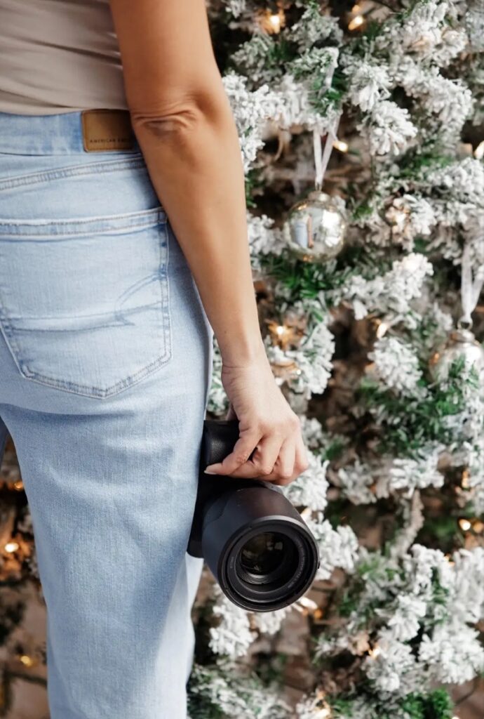 Branding photo of a woman holding a camera in front of a decorated Christmas tree, capturing holiday moments with a festive backdrop.