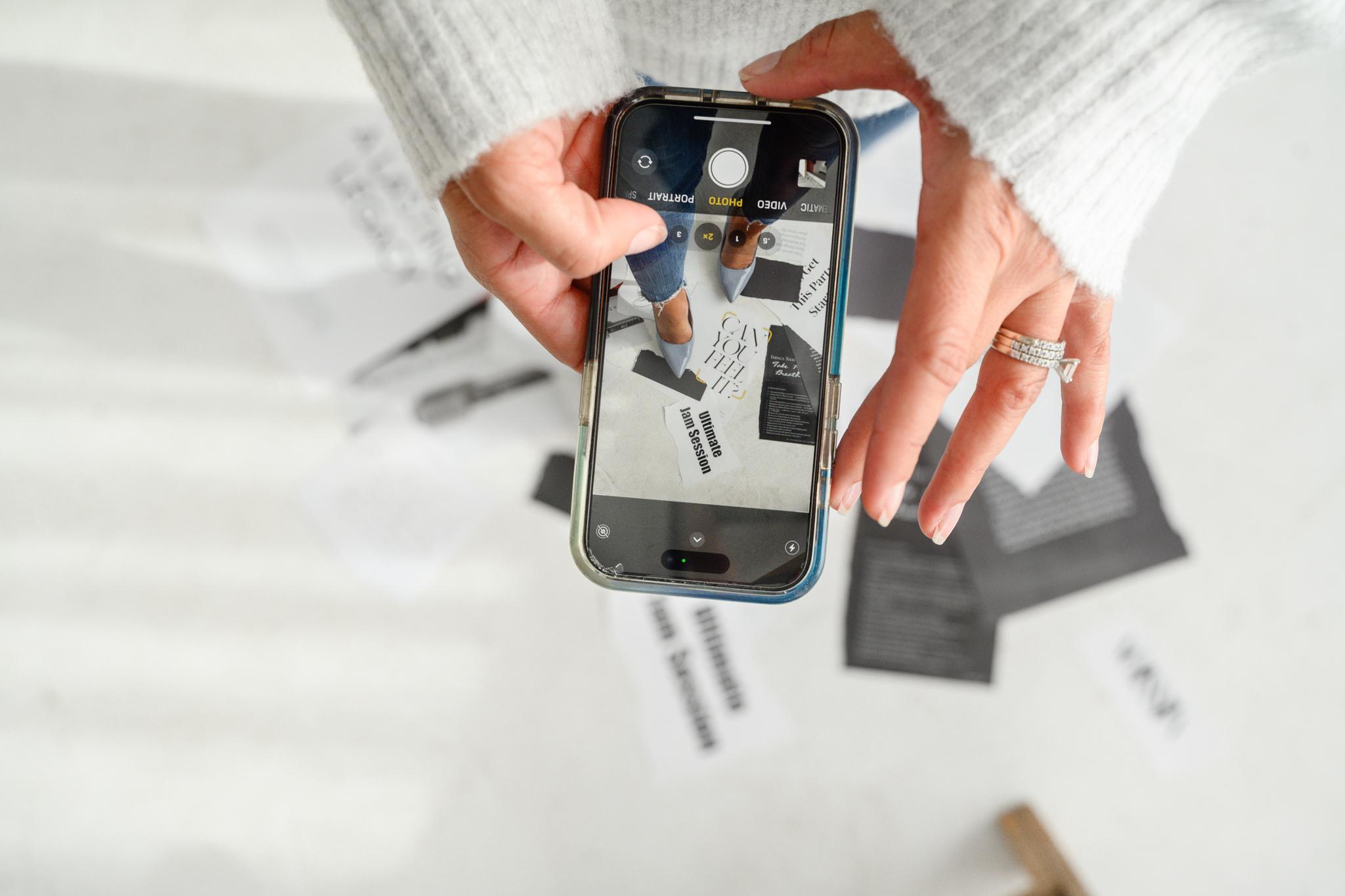 A woman holding a cell phone, viewing papers on the floor through the screen, showcasing images captured on her device.